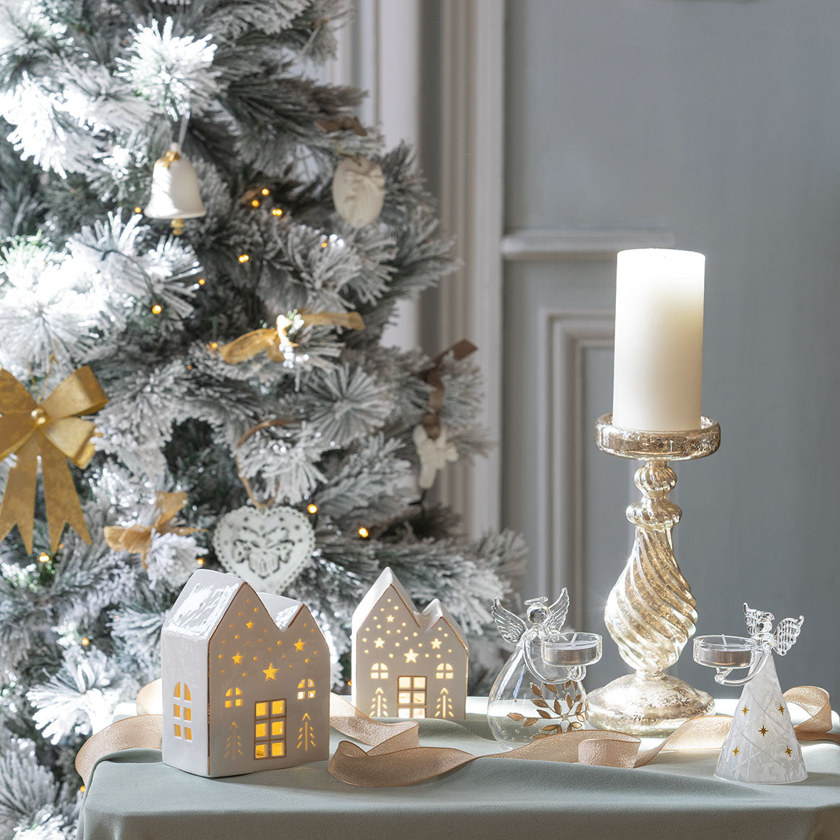 Photo mise en scène de 2 maisons décoratives de 2 tailles différentes posé sur une table, avec 1 ange porte-bougie en verre givré et un porte-bougie éclat d'ange. Un sapin de Noël en arrière plan