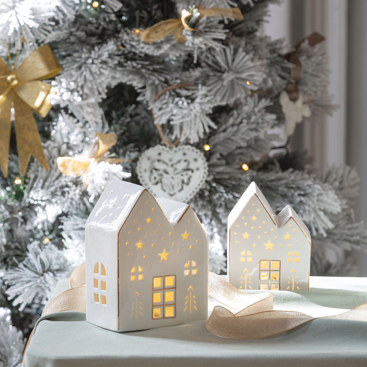 Photo mise en scène de 2 maisons décoratives de 2 tailles différentes posé sur une table et avec un sapin de Noël en arrière plan